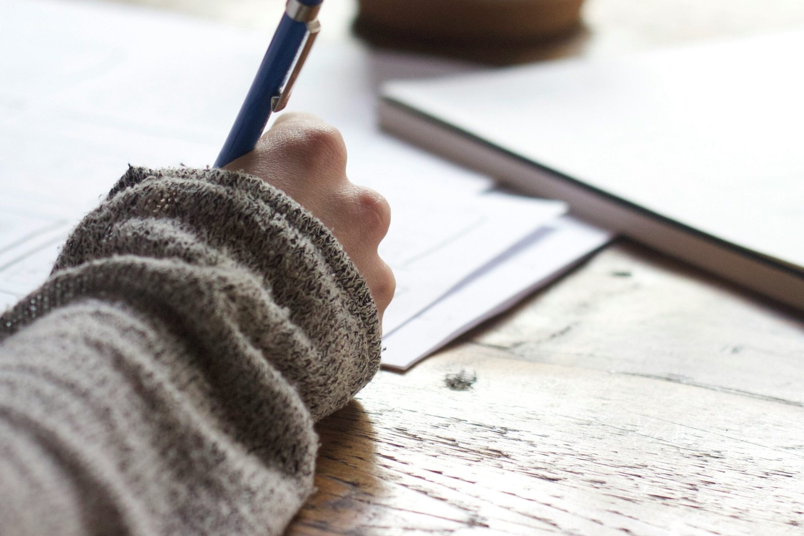 person writing on brown wooden table near white ceramic mug